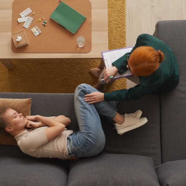 Depressed woman visiting psychologist High angle view of young woman lying on sofa in depression while psychologist talking to her at office