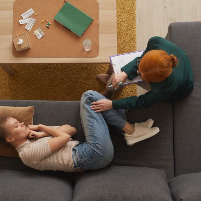 High angle view of young woman lying on sofa in depression while psychologist talking to her at office