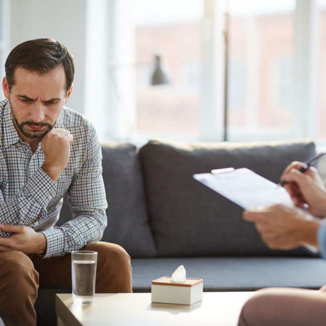 Pensive or stressed man sitting on couch in front of counselor and trying to explain his matter at individual session